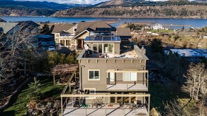 A modern two-story house with a large deck and solar panels on the roof, surrounded by a scenic lake and mountains. - Hazel House - Gorgeous, Views, Perfect Hood River Getaway! (Hood River)