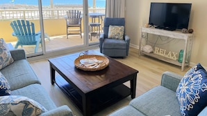 Ein Wohnzimmer mit Blick auf den Strand, ein blauer Sessel, ein Couchtisch aus Holz und ein Fernseher auf einer weißen Konsole.