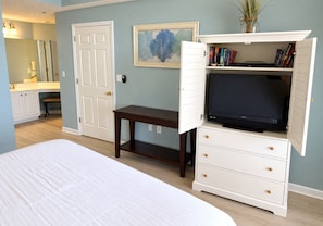 Un dormitorio con cama, televisor sobre un mueble, una mesita, un banco, una puerta y una estantería llena de libros.