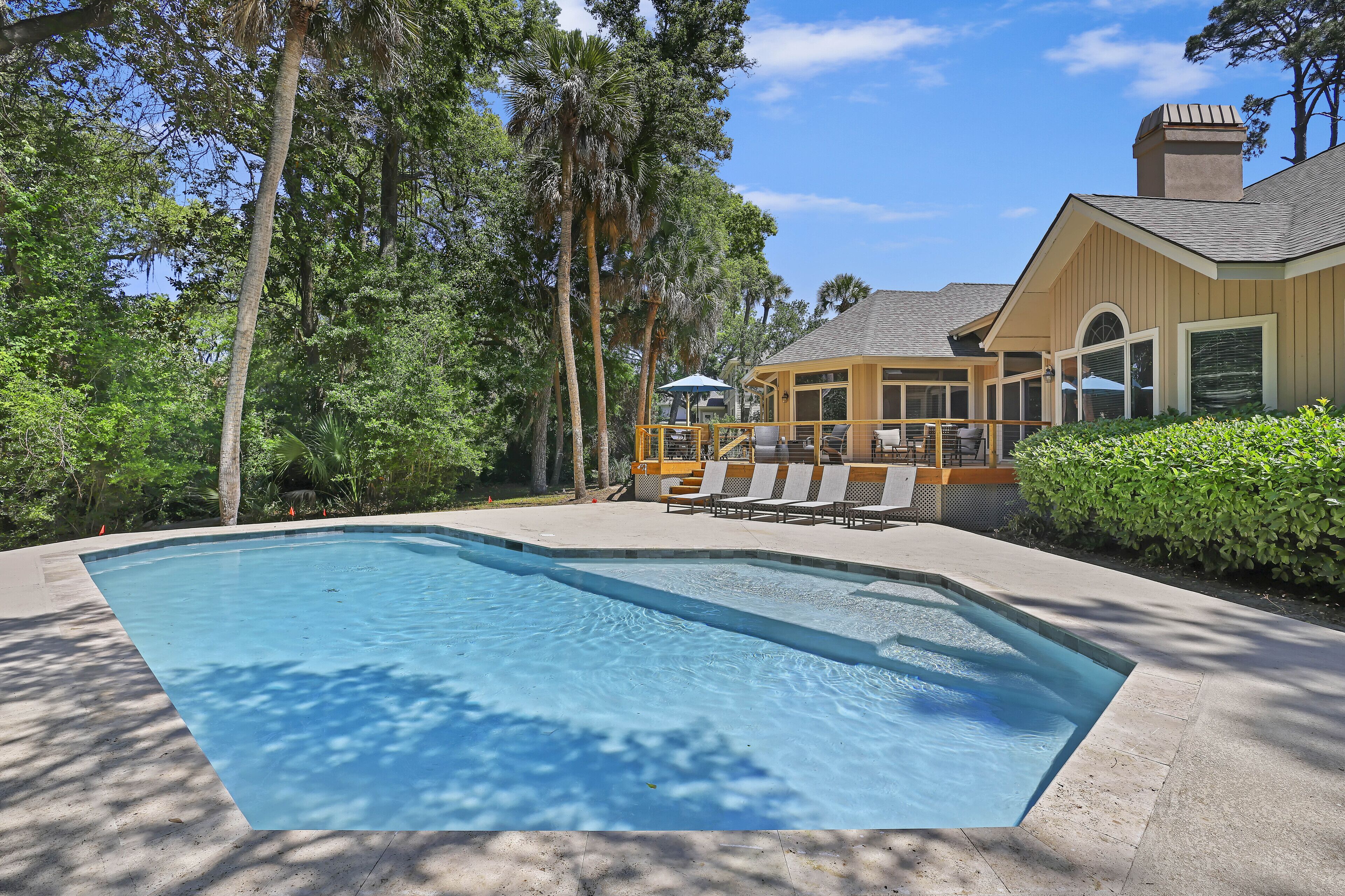 Une piscine avec des chaises longues et un patio couvert, le tout entouré d’arbres et d’une maison.