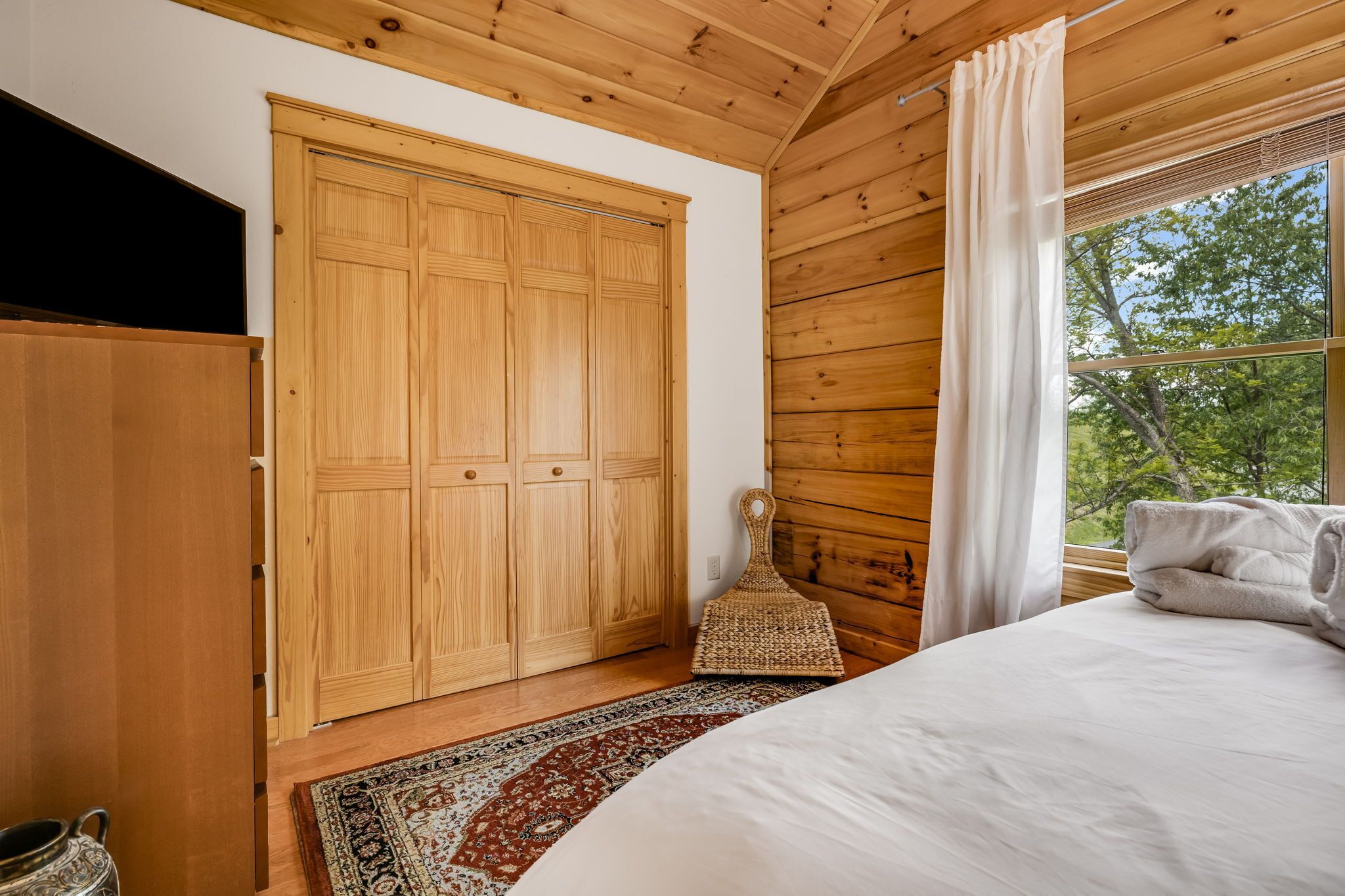 Un dormitorio con un armario grande de madera, una cama con sábanas blancas, un tapete estampado y una ventana con vistas a árboles.