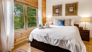 A wooden-paneled bedroom with a large window, a bed with white bedding, two framed pictures, and a bedside table with a lamp. - Skylands by Carolina Mornings | Hot Tub + Biltmore Pass 8 mins to Hatley Pointe (Mars Hill)