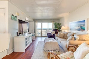 Een ruime woonkamer met een groot raam met uitzicht op de oceaan, een gezellige zithoek met een bank en fauteuils, een televisie en een klein keukentje.
