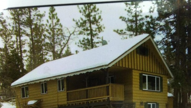 A two-story wooden cabin with a balcony, surrounded by snow and trees.