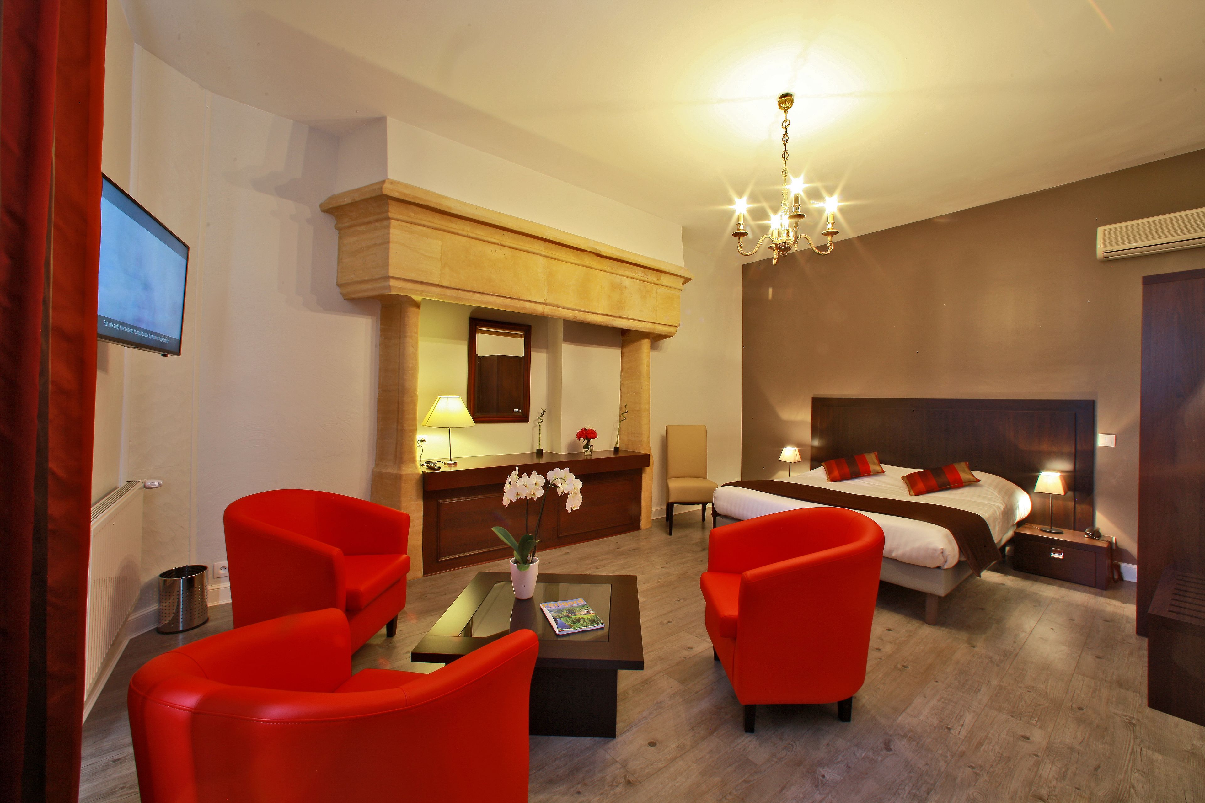 Une chambre d’hôtel avec un lit, deux fauteuils rouges, un bureau avec une lampe, une petite table avec un vase de fleurs et un téléviseur fixé au mur.