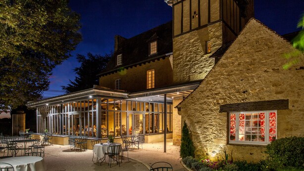 Un bâtiment traditionnel à la façade en pierre, doté d’un patio vitré avec des espaces de repos en plein air et d’une salle à manger intérieure visible.