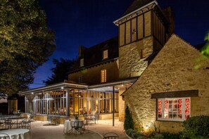 Un edificio tradicional con fachada de piedra, un patio cubierto de vidrio con mesas al aire libre, y un espacio interior comedor visible.