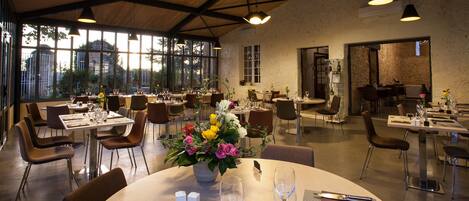 A spacious dining area with multiple tables and chairs, large windows offering a view of the outdoors, and a rustic wooden ceiling.
