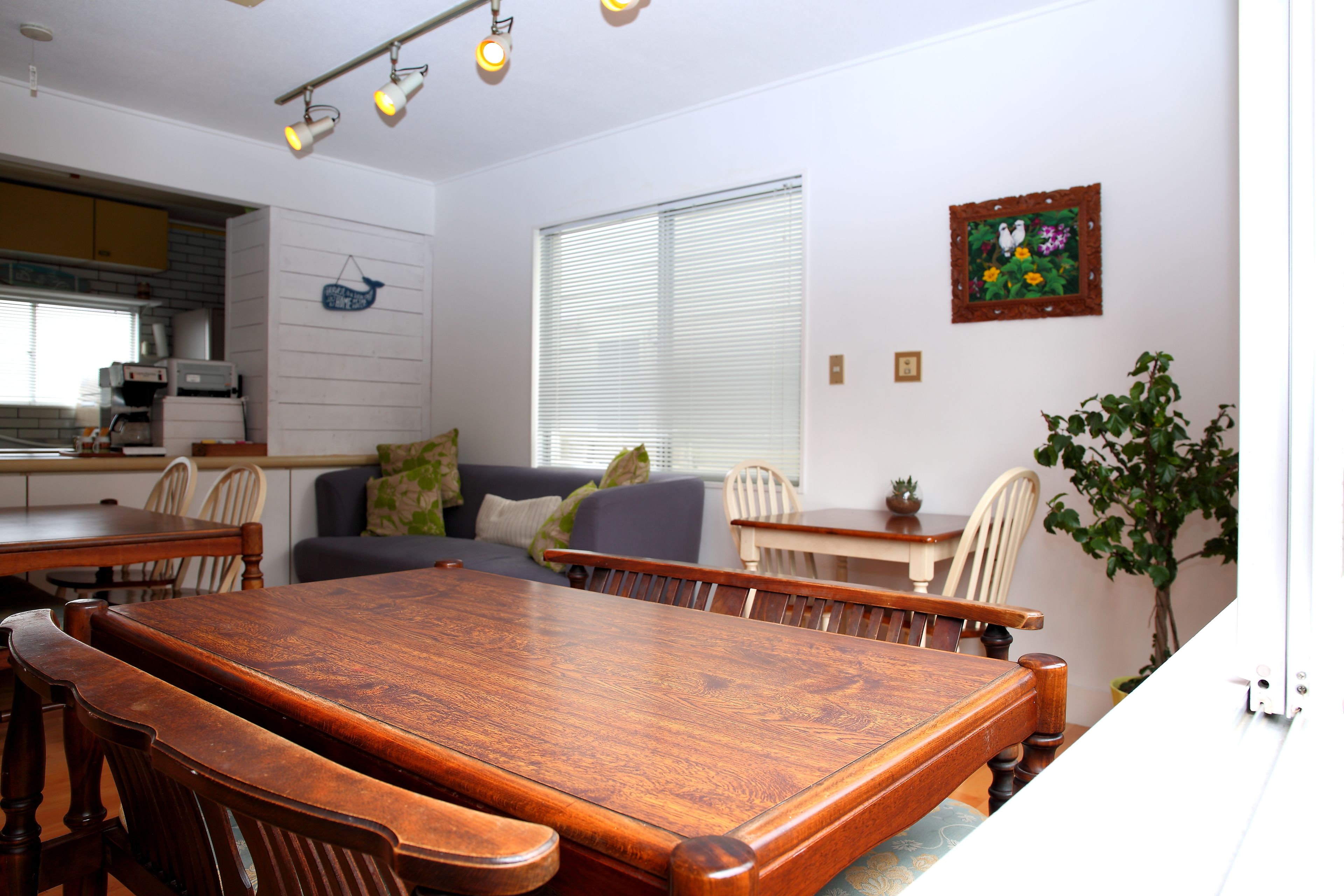 A dining area with a wooden table and chairs, a sofa, a window with blinds, and a kitchen in the background.