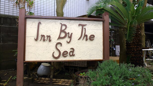 Una señal para 'Inn By The Sea' con un diseño rústico, rodeada de follaje y una palmera.