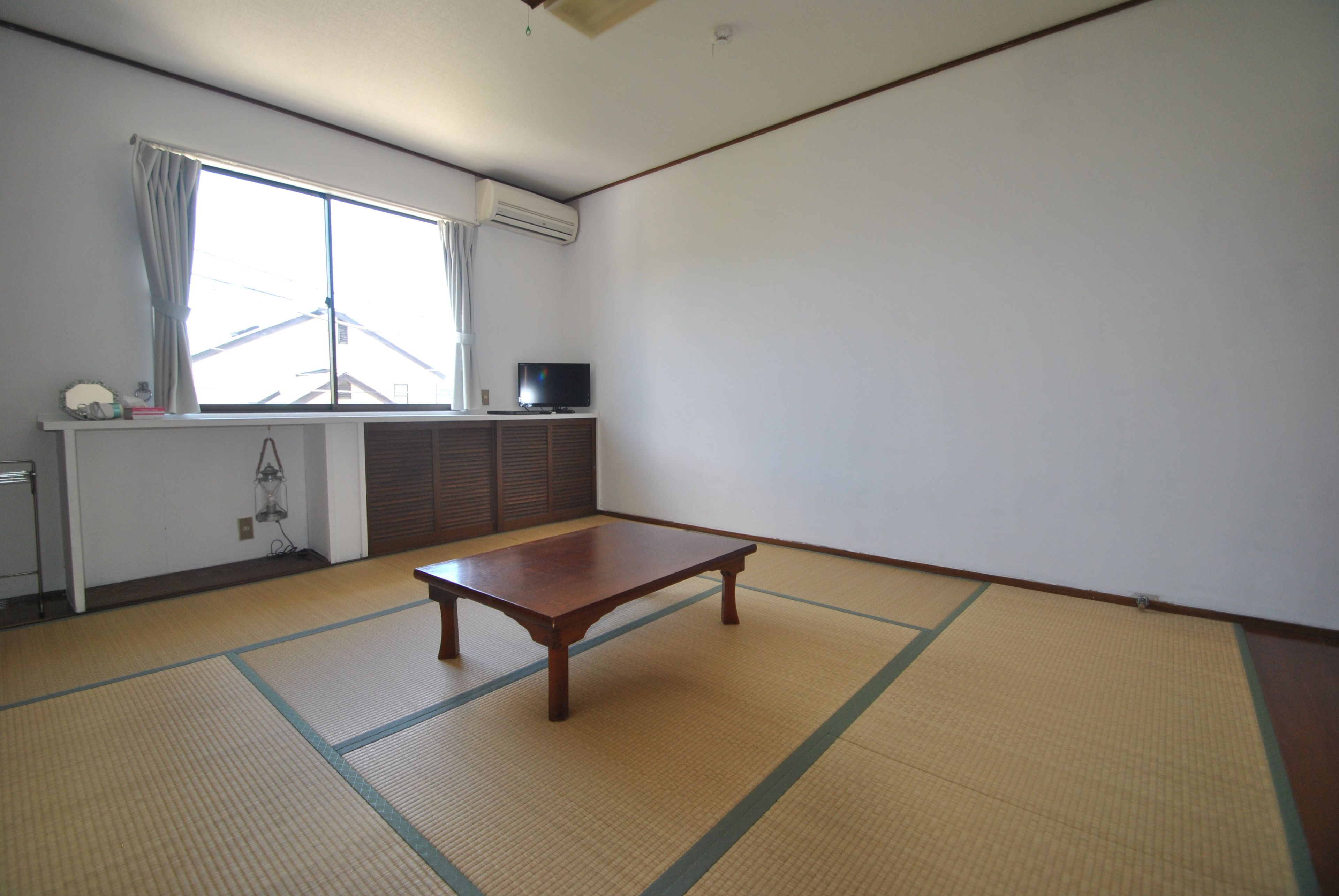 Una habitación con suelo de tatami, una mesa de madera, un televisor sobre un mueble y una ventana con cortinas.