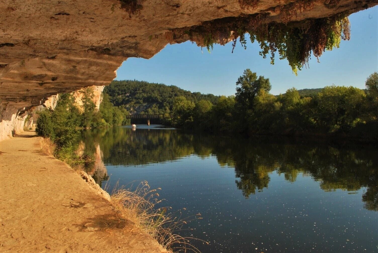 A serene river scene with a bridge, a sandy path, and lush greenery.