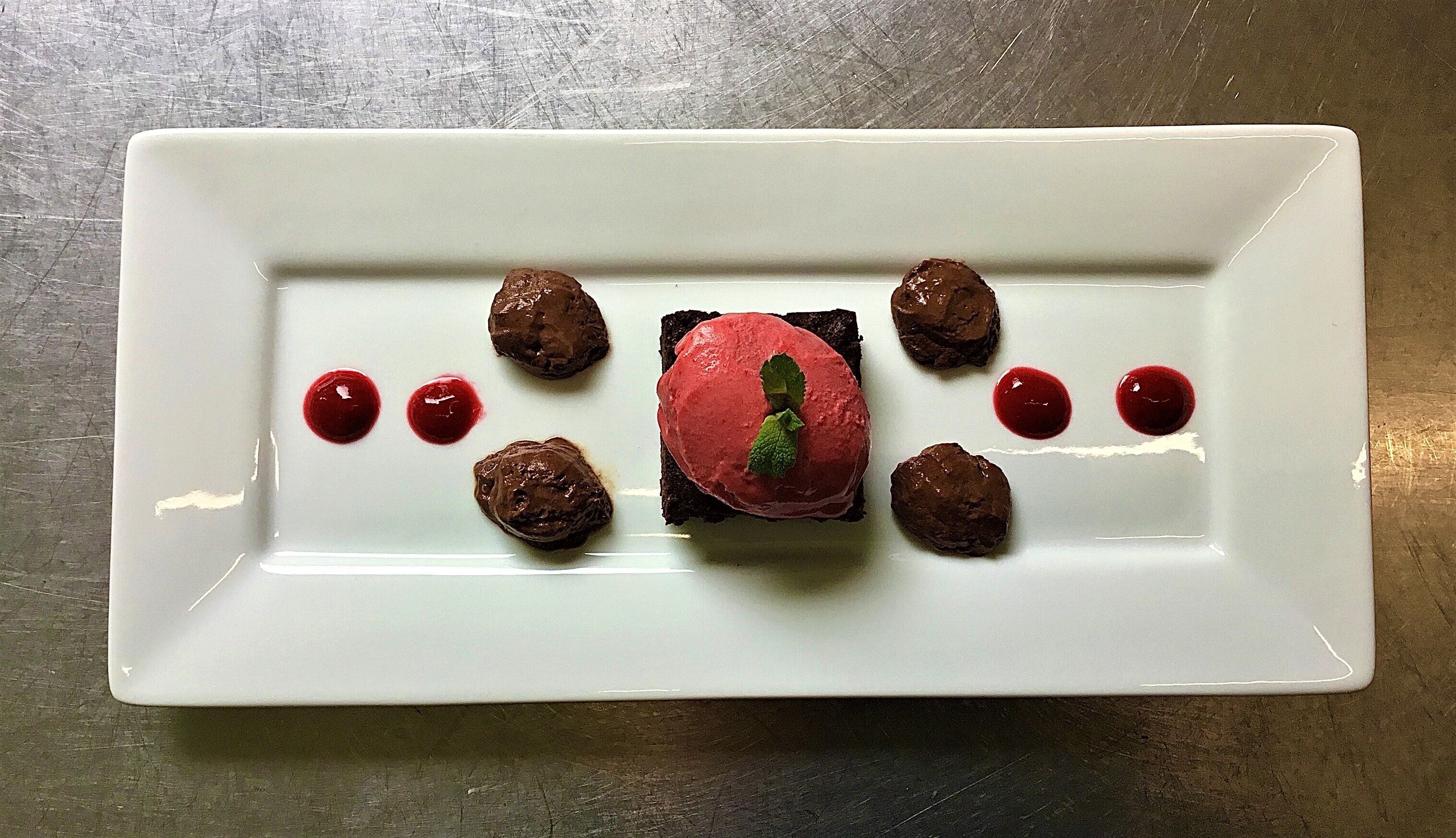 En rektangulær hvid tallerken med dessert bestående af en rød kugle, chokoladebolde og røde prikker.
