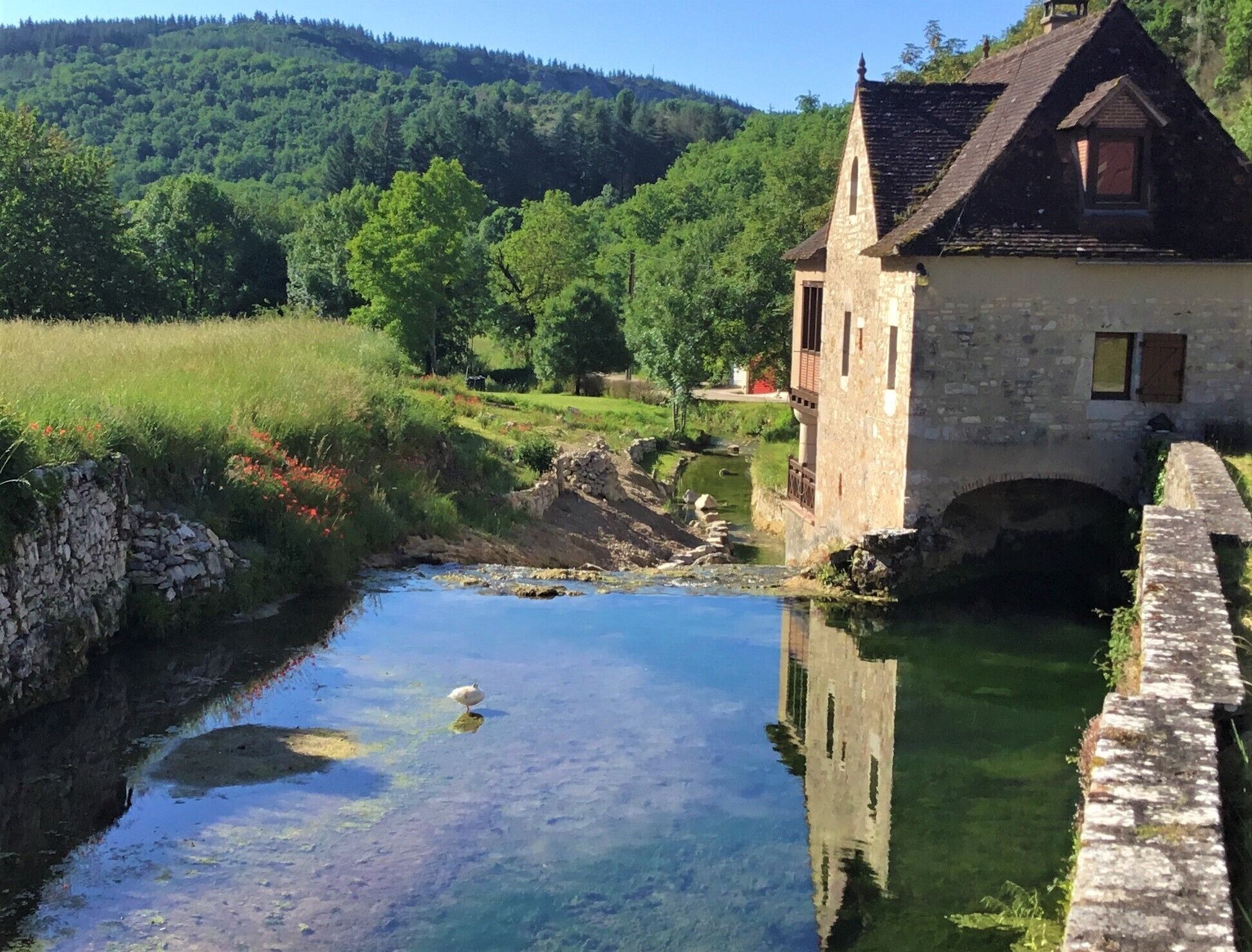 A stone building with a small bridge over a clear water stream, surrounded by lush greenery and hills.