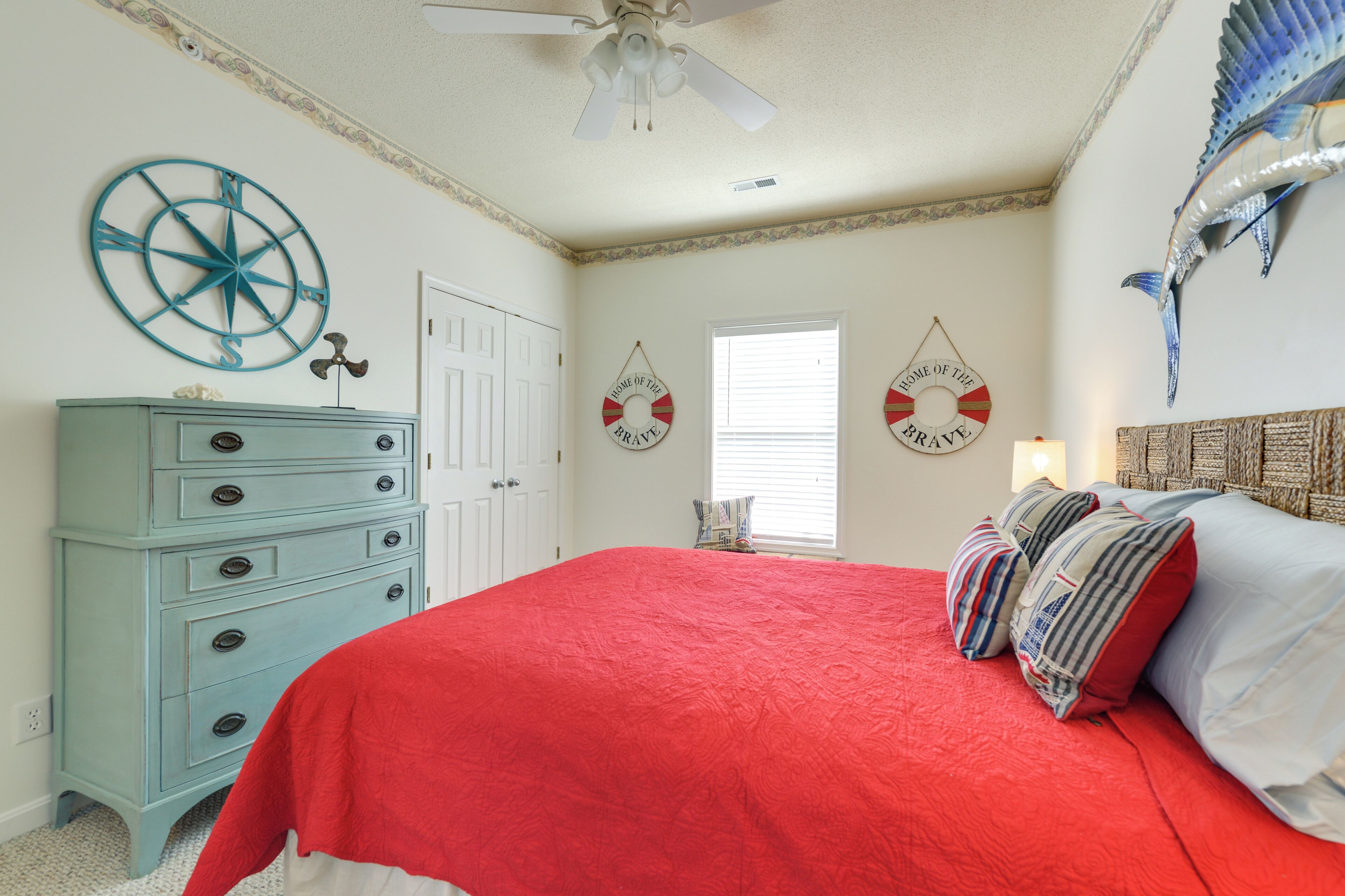 Une chambre à coucher avec une couvre-lit rouge, une commode bleue, un ventilateur de plafond et des décorations sur le thème de la marine.
