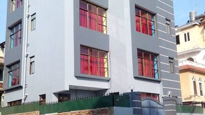 A modern multi-story building with a flat facade, large windows with red curtains, and a green fence at the base. - Nana Homes (Kathmandu)