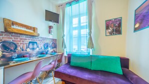 Un espace de vie moderne avec un mur en briques, une étagère en bois avec des verres, un canapé violet avec un coussin vert et un bureau avec un ordinateur et des chaises.