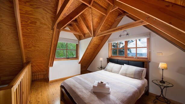 Une chambre avec un plafond en pente, des poutres en bois, un lit avec une literie blanche, deux tables de chevet avec des lampes et des fenêtres donnant sur un paysage verdoyant.