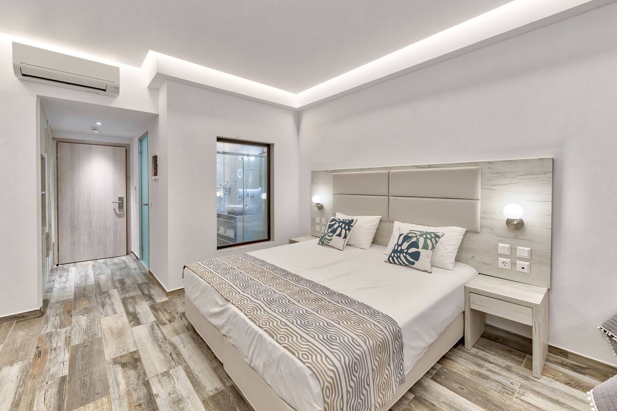 A modern bedroom with a large bed, a headboard with integrated lighting, a bedside table, and a view of the city through a large window.