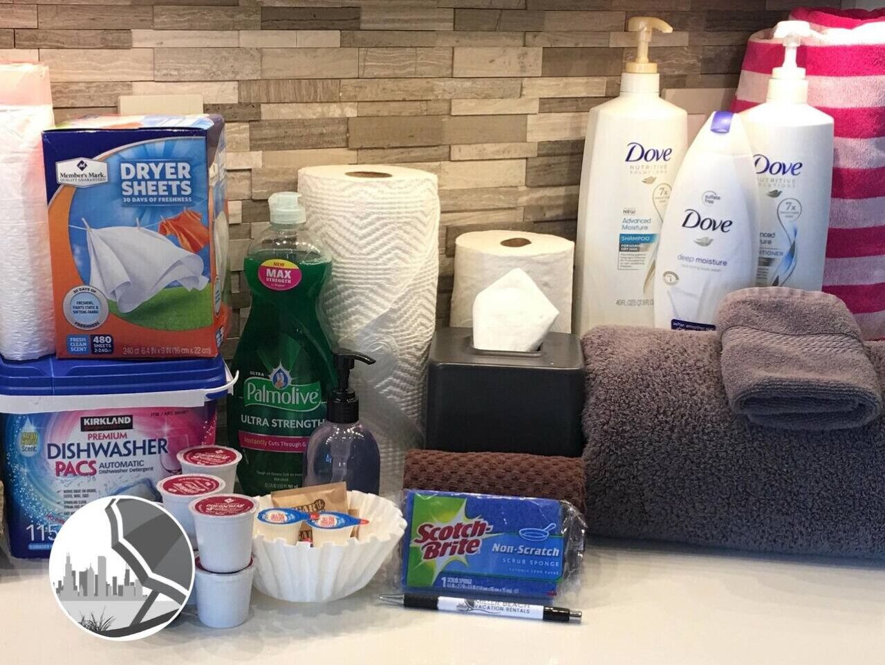 Un baño con salpicadero de azulejos, un montón de toallitas suavizantes, una botella de detergente Palmolive Ultra Fuerza, una caja de esponja Scotch-Brite No Rayar, una barra de jabón, una botella de loción Dove y una toalla enrollada.