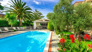 A swimming pool with lounge chairs, a covered seating area, and lush greenery. - Luxury Villa Fotini (Thasos)