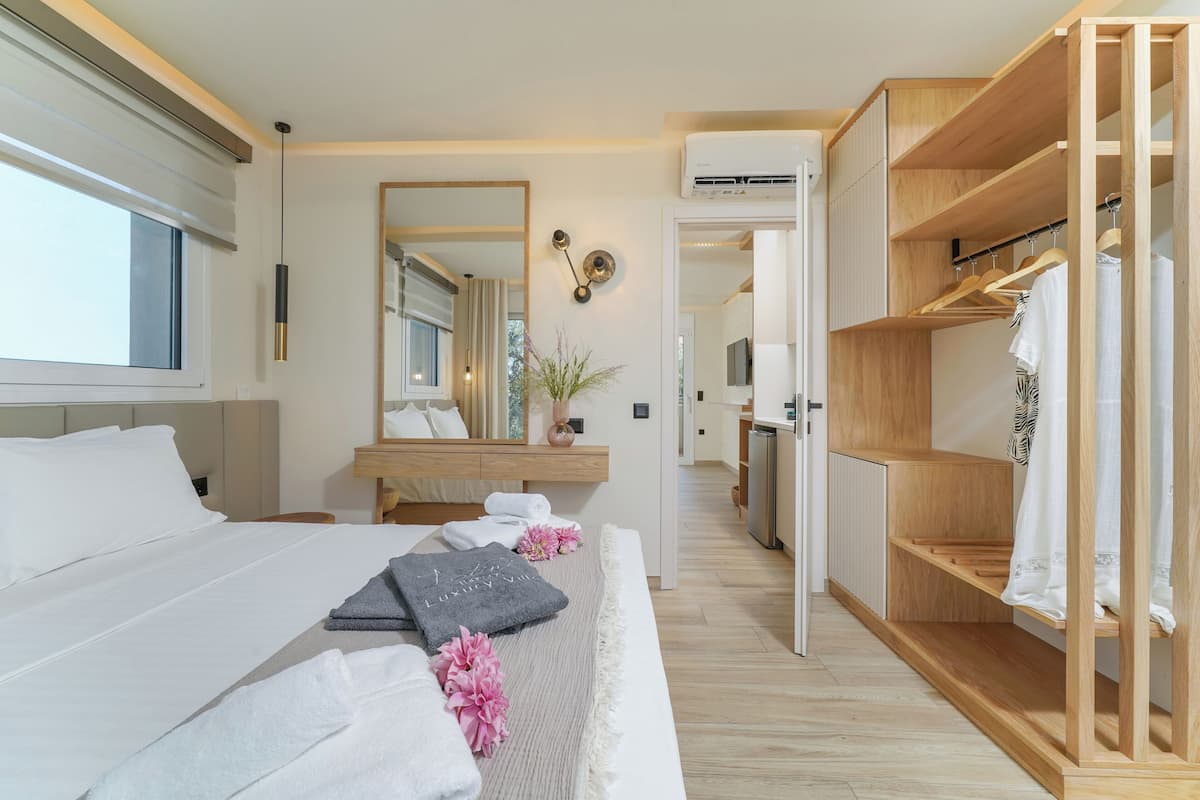 A modern hotel room with a large bed, a wooden wardrobe, a vanity with a mirror, and a view into the kitchen.