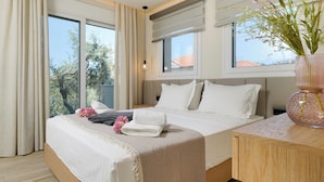 A hotel room with a large bed, a wooden nightstand, a vase with flowers, and a view of trees and buildings through the windows. - Luxury Villa Fotini (Thasos)