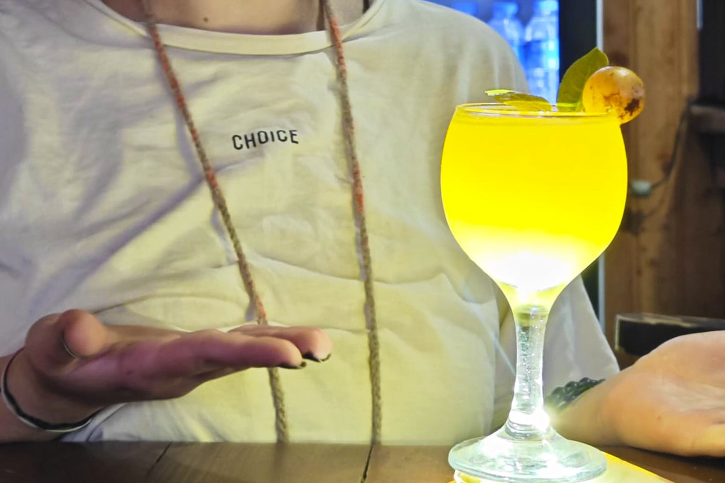 A person wearing a white shirt with the word "CHOICE" on it, sitting at a table with a yellow drink in a glass.