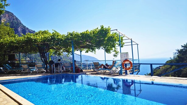 A clear blue pool with lounge chairs and a lifebuoy, surrounded by trees and a mountainous backdrop. - Fullmoon Camping (Fethiye)