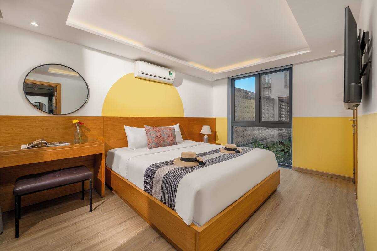 Una habitación de hotel moderna con una cama de madera, un escritorio, un banco, un espejo redondo, un equipo de aire acondicionado, un televisor y un ventanal con vistas.