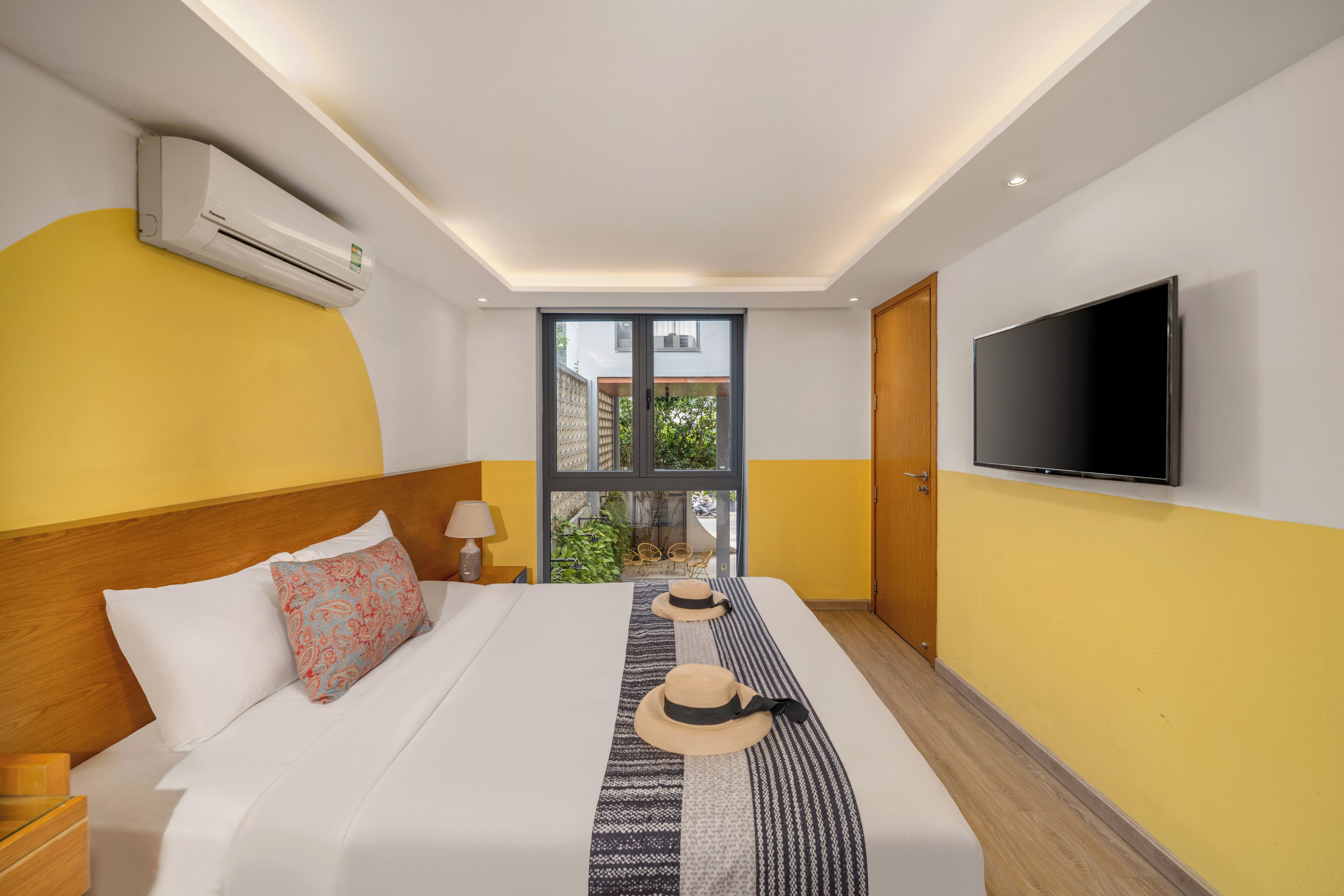 Habitación de hotel con cama, televisión, aire acondicionado y ventana con vista a follaje.