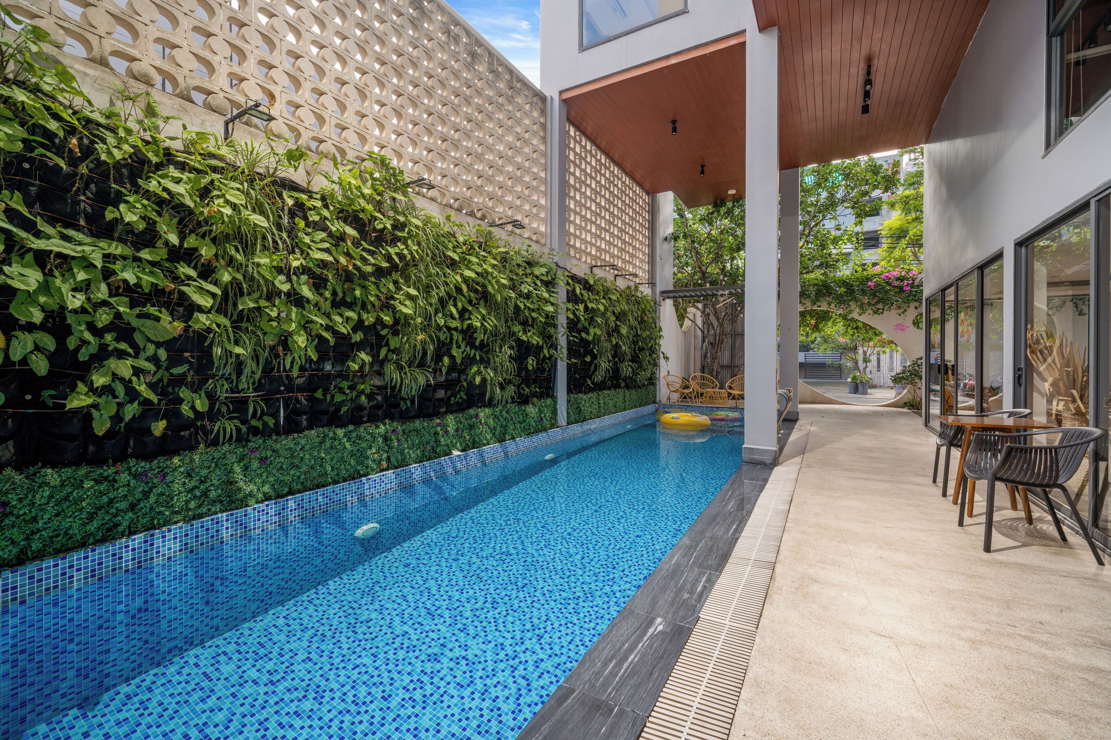 A modern outdoor pool area with a tiled blue pool, surrounded by lush greenery and a wooden pergola.