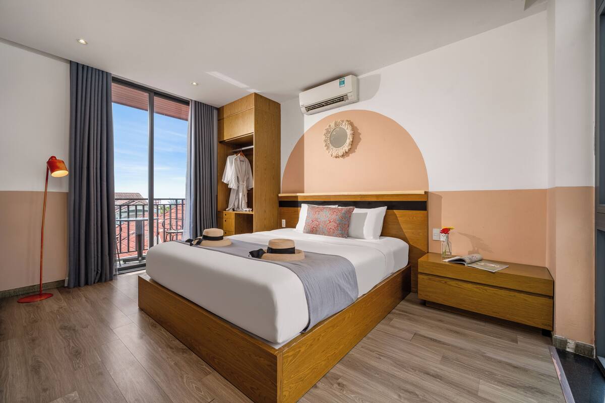 Habitación de hotel moderna con una cama grande, cabecera de madera, una mesita de noche, una lámpara de pie y vistas a la ciudad a través de ventanas amplias.