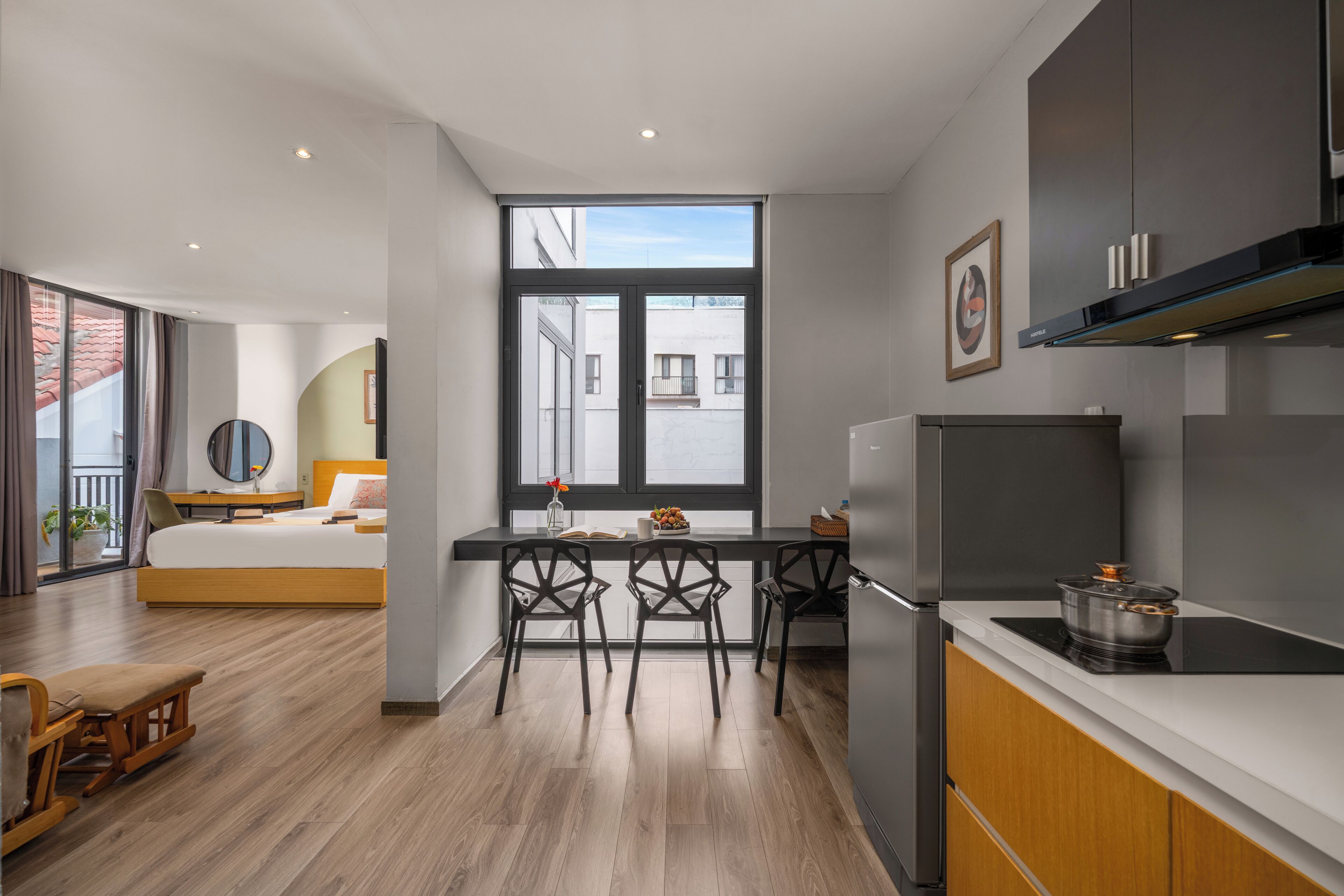 Ein modernes Hotelzimmer mit einem Bett, einem Essbereich mit Stühlen, einer Küche inklusive Kühlschrank und Herd sowie einem großen Fenster mit Blick auf die Straße.