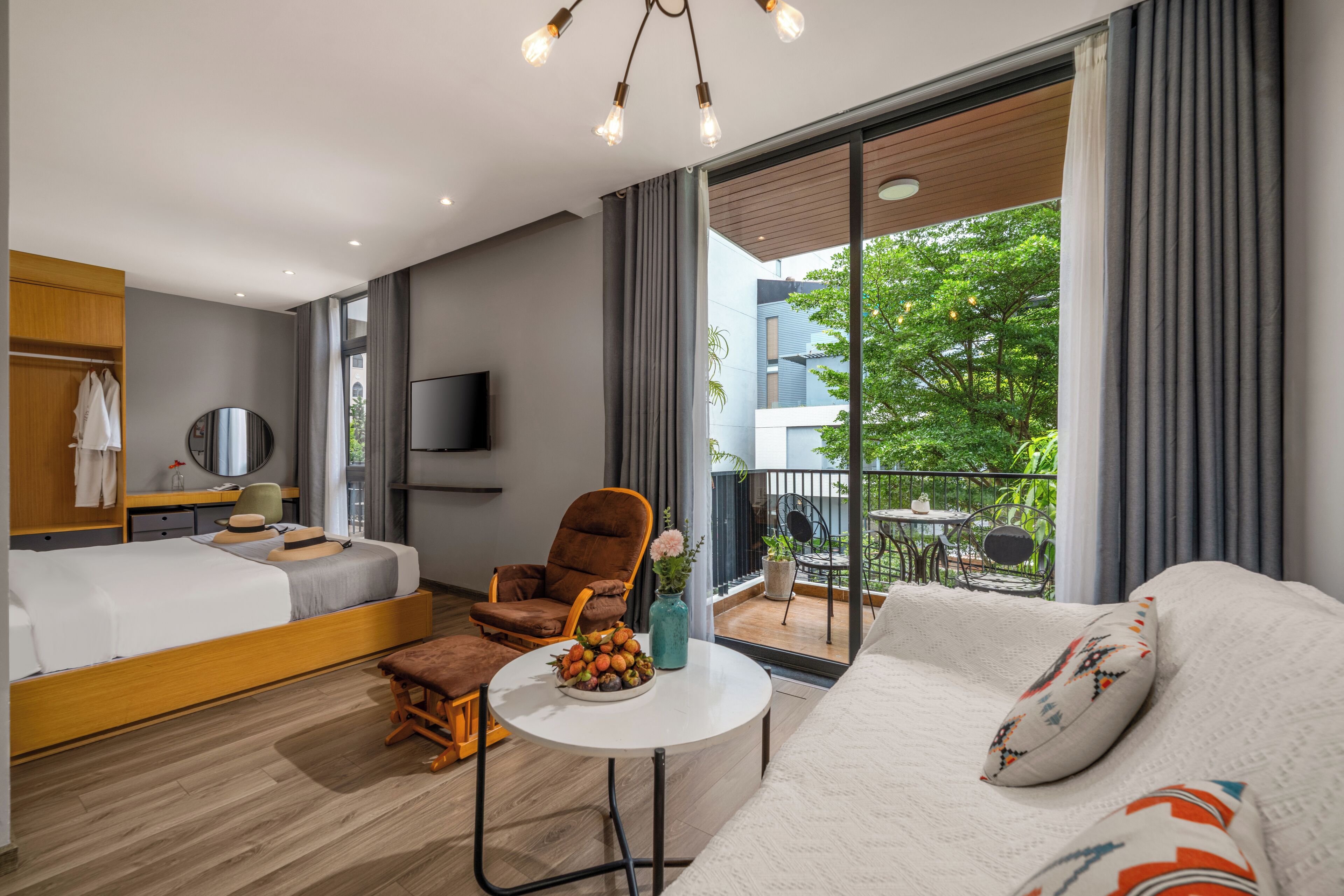 A modern hotel room with a bed, sofa, armchair, and a small table with fruit.