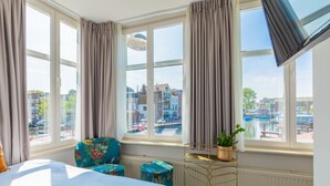 A hotel room with a large bed, a window with curtains, a view of buildings and a canal, a small table with a plant, and a colorful chair. - LOT Hotel (Leiden)