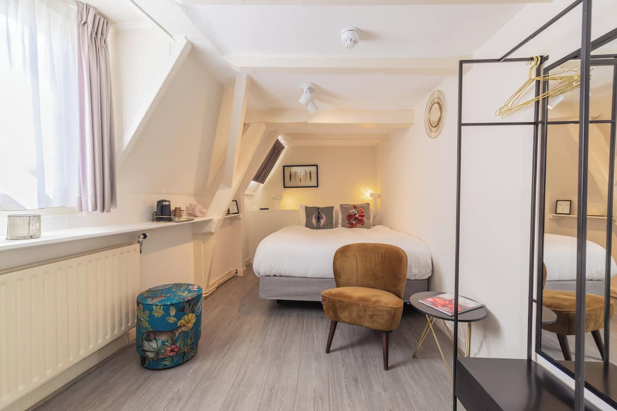 A bedroom with a bed, a chair, a small table, a radiator, a window with curtains, and a mirror.