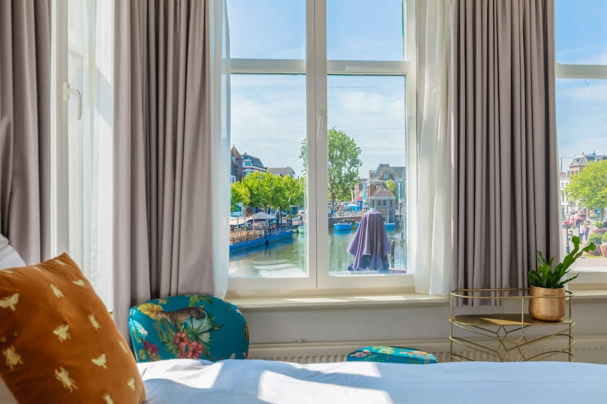 A hotel room with a large window offering a view of a canal and buildings. There is a bed with a blue pillow and a small table with a plant.