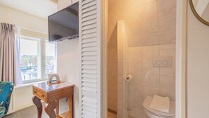 A bathroom with a toilet, a shower with a glass door, a wooden desk with a chair, and a TV mounted above the desk. - LOT Hotel (Leiden)