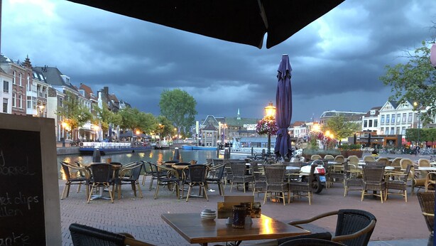 An outdoor café with wicker chairs and tables, overlooking a canal with boats and historic buildings in the background. - LOT Hotel (Leiden)