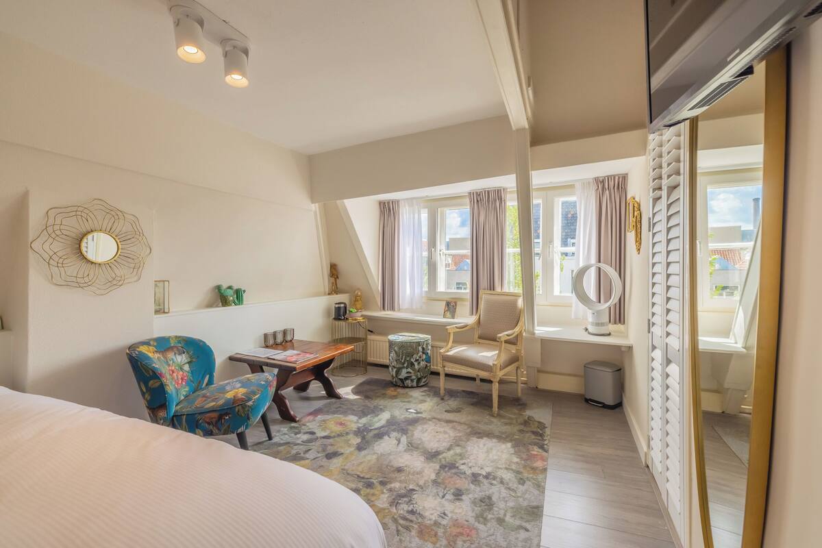 A bedroom with a bed, a chair, a table, a mirror, and a window with curtains.