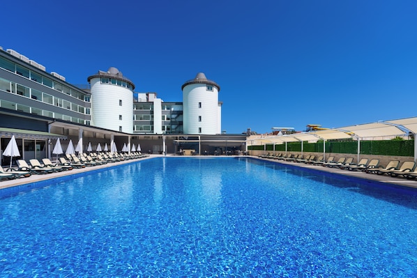 Una gran piscina con sillones de descanso y sombrillas, rodeada de edificios modernos con torres cilíndricas.
