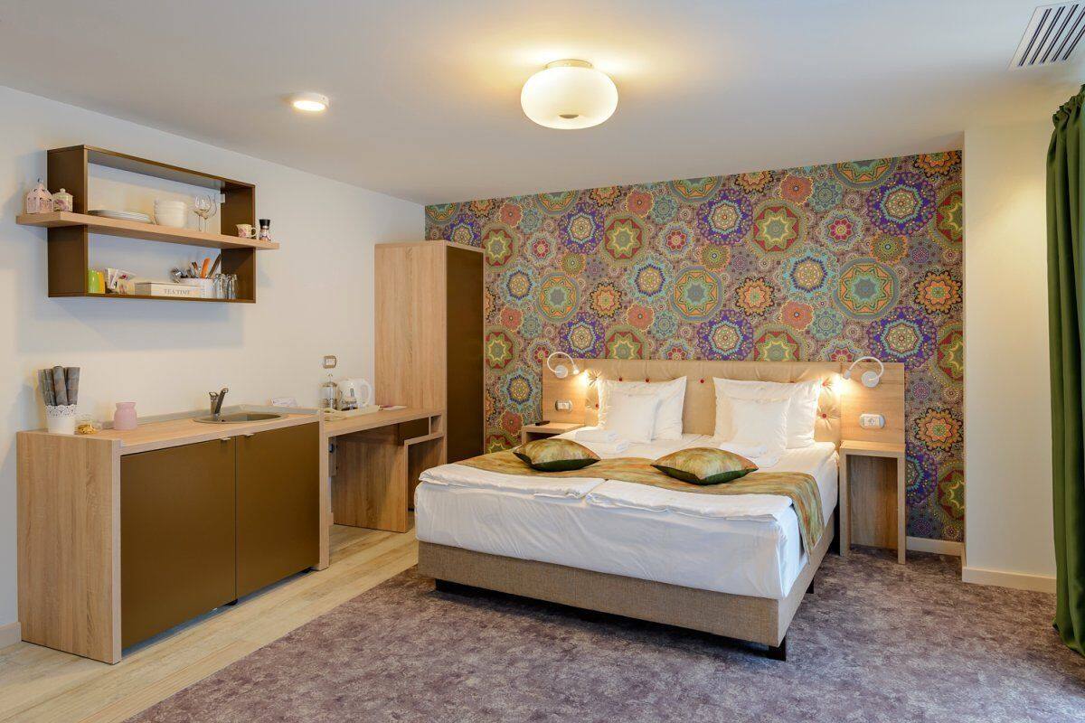 A compact hotel room with a bed, bedside tables, a built-in desk, and a kitchenette.