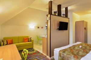 Habitación de hotel compacta con un sofá verde, una cama con manta estampada, un escritorio pequeño con una silla y un televisor montado en la pared.