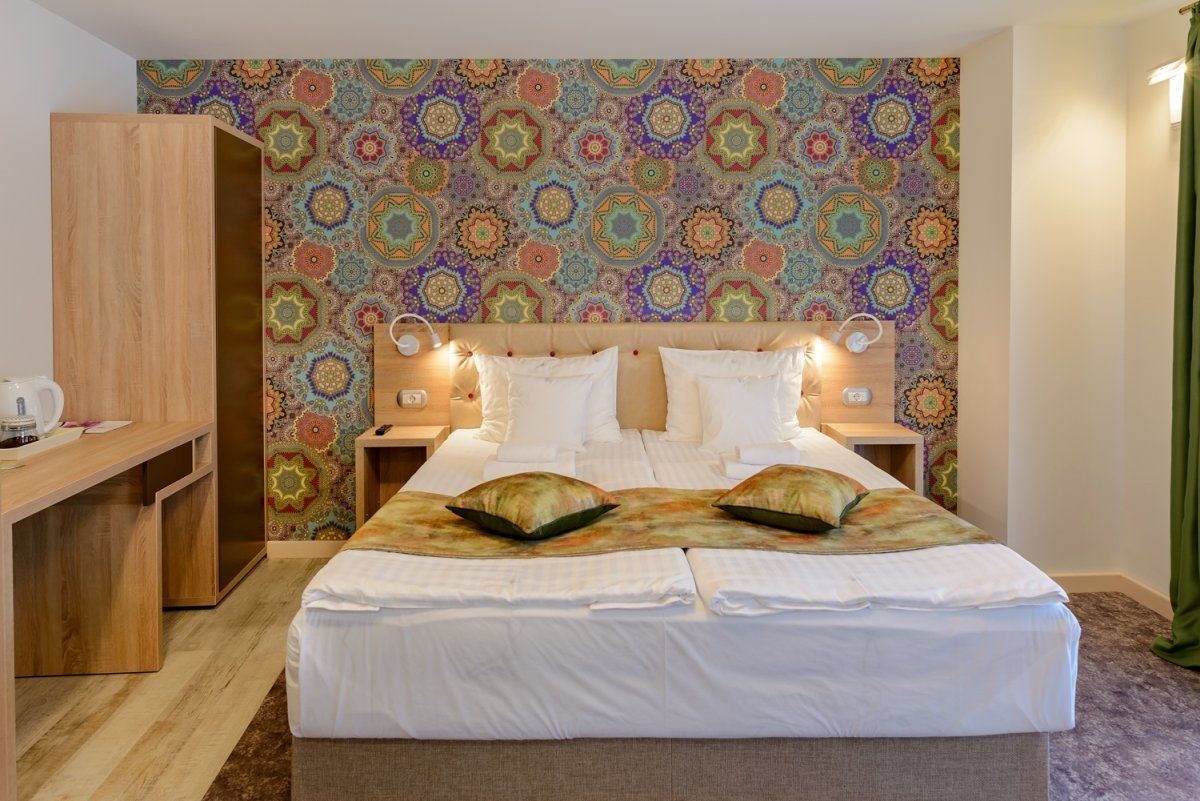 Una cama doble con sábanas blancas y dos almohadas decorativas, un escritorio de madera y una pared con un motivo geométrico colorido.