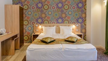 Una cama doble con sábanas blancas y dos almohadas decorativas, un escritorio de madera y una pared con un motivo geométrico colorido.