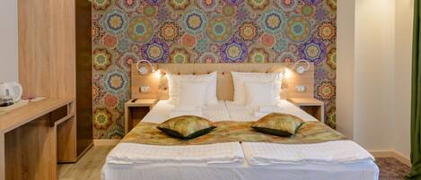 Una cama doble con sábanas blancas y dos almohadas decorativas, un escritorio de madera y una pared con un motivo geométrico colorido.