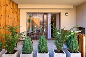 Un área de estar moderna al aire libre con un ventanal de vidrio que da a una habitación, rodeada de plantas perennes en macetas.