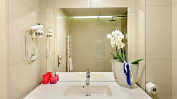 A bathroom with a white sink, a mirror, a shower, and a vase with white flowers.