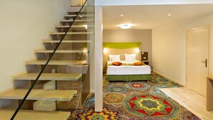 A modern hotel room with a staircase leading to a balcony, a bed with a green headboard, and a colorful patterned carpet. - Blumenhaus Sibiu (Sibiu)